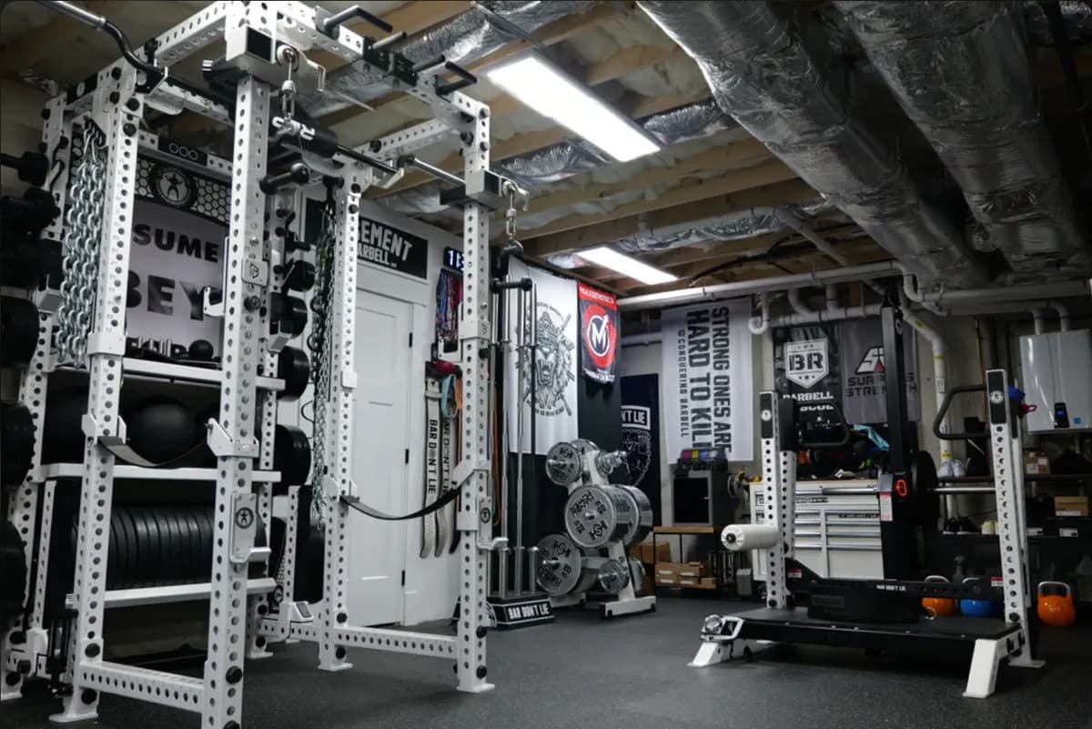 Photograph of Basement Barbell: a white six-post power rack hung with chains and bands, black plates stored on the uprights, banners on the walls and exposed silver ductwork on the ceiling